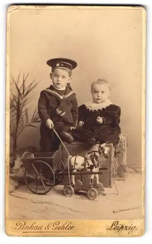 Fotografie Pinkau & Gehler, Leipzig, Portrait zwei kleine Kinder mit Spielzuegpferd samt Karren und Peitsche