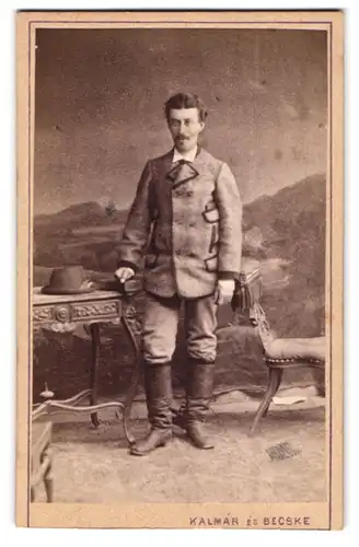 Fotografie Kalmar es Becske, Becsböl, Portrait junger Mann im Anzug mit Lederstifel und Hut auf dem Tisch