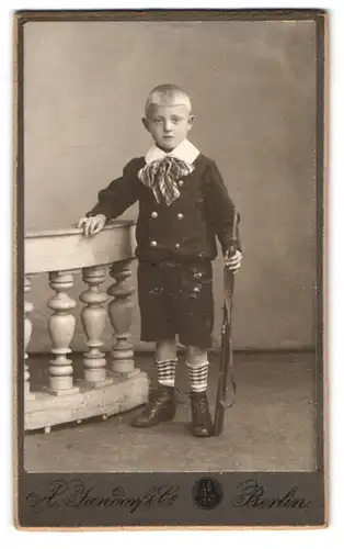 Fotografie A. Jandorf & Co., Berlin, Portrait niedlicher Knabe im Anzug mit Spielzeug Gewehr im Atelier