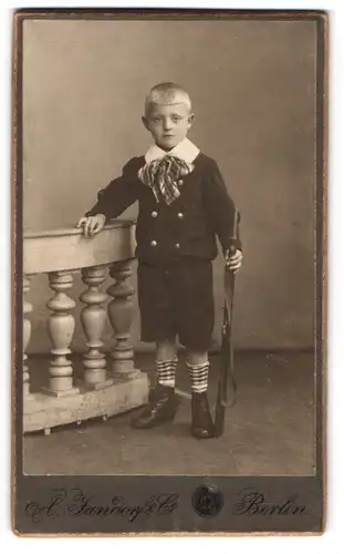 Fotografie A. Jandorf & Co., Berlin, Portrait junger Knabe im Anzug mit kurzer Hose und Gewehr bei Fuss