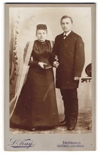 Fotografie L. Kny, Ebersbach i. Sa., Portrait Eheleute im schwarzen Hochzeitskleid und Anzug mit Schleier