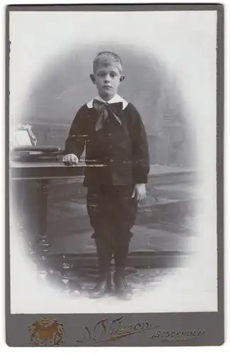 Fotografie N. Nilsson, Stockholm, Wasagatan 9, Junge mit traurigem Blick und Segelohren im Anzug mit kurzen Hosen