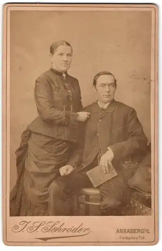 Fotografie J. S. Schröder, Annaberg i. S., Carlsplatz 756, Portrait Dame im schwarzen Kleid und Herr mit Buch