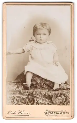 Fotografie Joh. Heine, Georgswalde, Kleinkind im kurzen Kleidchen auf einem Sitzmöbel