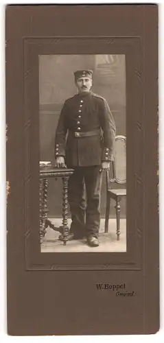 Fotografie W. Boppel, Gmünd, Soldat in Uniform mit Krätzchen an einen Tisch lehnend