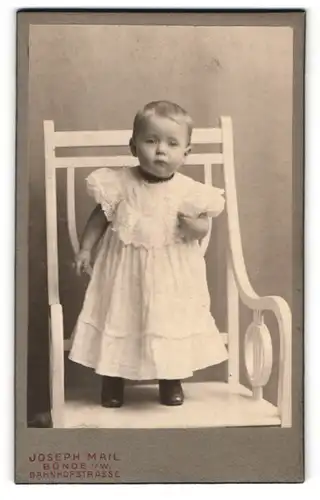 Fotografie Joseph Mail, Bünde /Westf., Bahnhofstrasse, Kind im Taufkleid stehend auf einem Stuhl