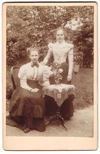 Fotografie unbekannter Fotograf und Ort, Zwei junge Frauen in taillierten Kleidern an einem Tischchen im Garten