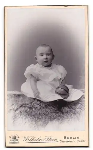Fotografie Wilhelm Stein, Berlin, Chausseestr. 65-66, Süsses Kleinkind im Kleid mit Ball sitzt auf Fell