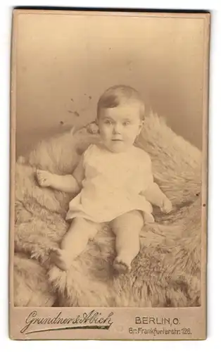Fotografie Grundner & Abich, Berlin, Gr. Frankfurterstr. 126, Portrait süsses Baby im Hemdchen auf Fell sitzend