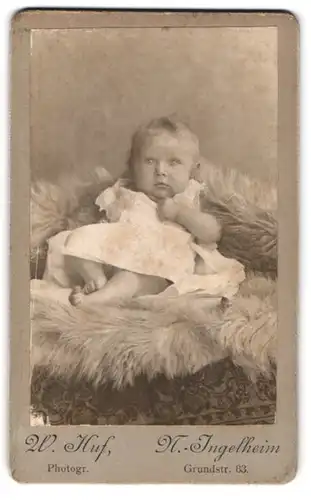 Fotografie W. Huf, N.-Ingelheim, Grundstr. 83, Portrait süsses blondes Baby im Kleidchen auf Fell liegend