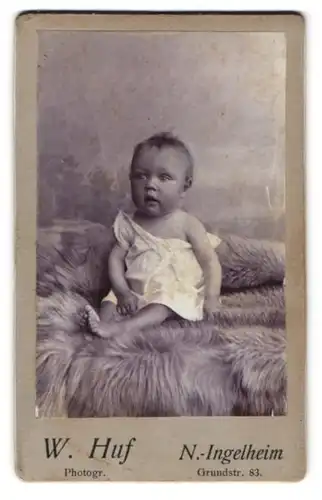 Fotografie W. Huf, N.-Ingelheim, Grundstr. 83, Portrait blondes niedliches Baby im Hemdchen auf Fell sitzend