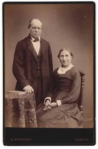 Fotografie H. Schröder, Lübeck, Betagtes Ehepaar neben Tisch