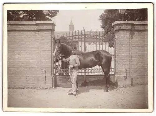 Fotografie unbekannter Fotograf und Ort, K.u.K. Soldat in Uniform mit seinem Pferd am Kasernentor