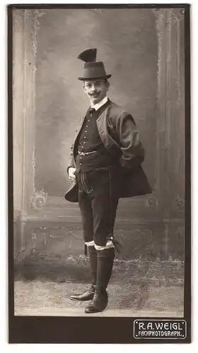 Fotografie R. A. Weigl, Ort unbekannt, Junger Mann mit Federhut in einer Tracht, Lederhosen
