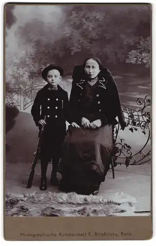 Fotografie E. Strassnicky, Enns, Mutter und Sohn in edler Tracht gekleidet, Knirps mit Karabiner