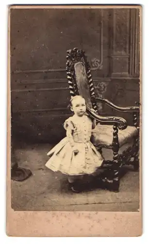 Fotografie Wells & Grey, Brighton, 144. Western Road, Kleinkind im weissen Kleidchen neben einem Holzstuhl posierend