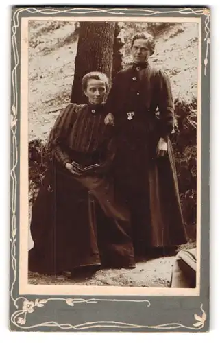 Fotografie unbekannter Fotograf und Ort, Zwei ernst blickende Damen in langen Kleidern vor einem Baumstamm