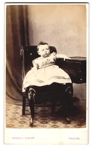 Fotografie Brown & Dunlop, Falkirk, Kleinkind im weissen Kleidchen auf einem Stuhl sitzend