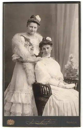 Fotografie J. Fuchs, Berlin, Portrait zwei Damen in weissen Kleidern mit Pelzstola und Haarschmuck