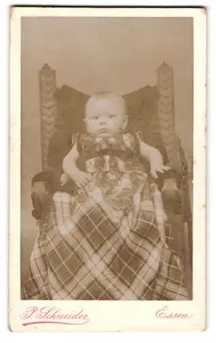 Fotografie P. Schneider, Essen, Kettwigerstrasse 38, Kleinkind im karierten, übergrossen Gewand in einem Sessel sitzend