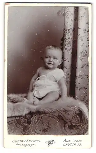Fotografie Gustav Kneisel, Aue /Erzgeb., Niedliches Kleinkind im weissen Gewand auf Pelz sitzend