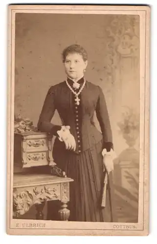Fotografie Ernst Ulbrich, Cottbus, Ernste junge Frau in tailliertem Kleid mit Faltenrock und grosser Kreuzkette