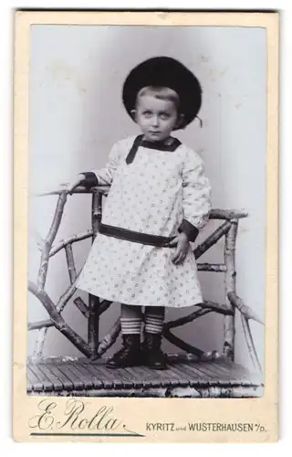 Fotografie E. Rolla, Kyritz, Kleinkind im weissen Kleidchen mit Pelzmütze auf einer Bank stehend