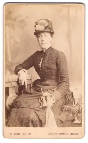 Fotografie Stuart Brothers, London, 47 Brompton Road, Junge Dame mit hohem Hut und Stockschirm im taillierten Kleid