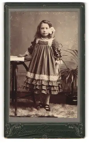 Fotografie Williams & Williams, Ort unbekannt, Kleines Mädchen mit Haarschleife in einem weiten Rüschenkleid