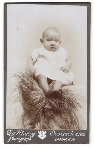 Fotografie Gg. Klersy, Oestrich a /Rh., Landstr. 18, Süsses Kleinkind im Hemd sitzt auf Fell