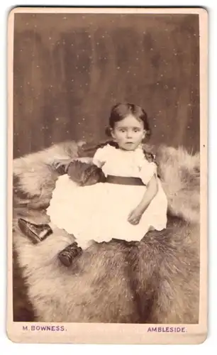 Fotografie M. Bowness, Ambleside, Kleines Mädchen im Kleid sitzt auf Fell