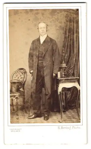 Fotografie H. Hering, London, 137 Regent Street, Herr im Anzug neben einem Tisch