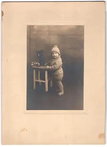 Fotografie unbekannter Fotograf und Ort, niedlicher kleiner Knabe in Winterkleidung mit seinem Teddy Bär auf dem Tisch