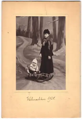 Fotografie unbekannter Fotograf und Ort, Mutter mit ihrem Kind im Winterkleid vor einer Winter Studiokulisse, Schlitten