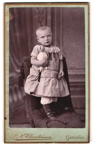 Fotografie L. N. Christensen, Haderslev, Apothekergade 310, Kind mit kurzen Haaren im Kleid mit Spielzeugfigur