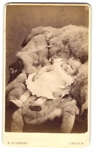 Fotografie R. Slingsby, Lincoln, 168, High Street, Süsses Kleinkind im Kleid liegt auf Fell