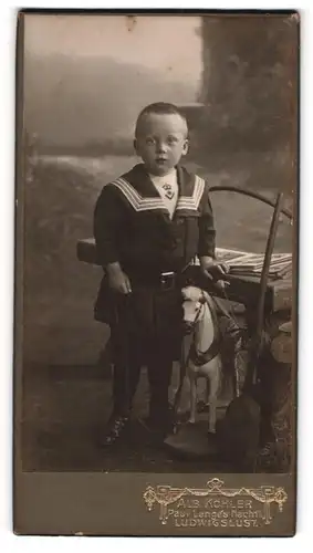 Fotografie Alb. Köhler, Ludwigslust, Portrait kleiner Junge im Matrosenkleid mit Spielzugpferd