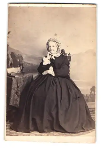 Fotografie unbekannter Fotograf und Ort, Grossmutter mit Haube im Sonntagskleid vor Studiokulisse sitzend