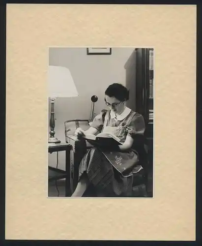 Fotografie unbekannter Fotograf und Ort, Hausfrau im karierten Kleid mit Schürze liest ein Buch um 1936