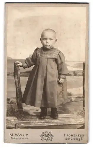 Fotografie M. Wolf, Pforzheim, Schulberg 3, Portrait süsses Kleinkind im Kleidchen auf einer Holzbank stehend
