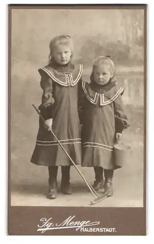 Fotografie Ig. Menge, Halberstadt, Zwei kleine Mädchen in identischen Kleidern mit Spielrechen und Eimer
