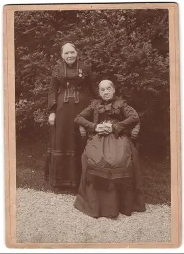 Fotografie unbekannter Fotograf und Ort, zwei ältere Damen mit Orden in Biedermeierkleidern posieren im Garten