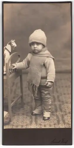 Fotografie Hanns Teich, Berlin, Portrait Kleinkind in Winterkleidung mit Schal und Mütze, Spielzeugpferd