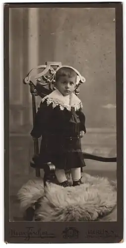 Fotografie Max Fischer, Berlin, Kleinkidn im schwarzen Samtkleid mit Spitzenkragen stehend auf einem Stuhl
