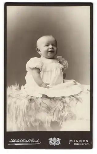 Fotografie Carl Beste, Minden, Bäckerstr., Strahlendes Kleinkind im weissen Kleid auf Pelz sitzend