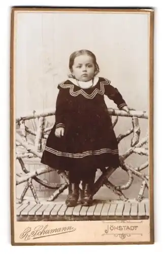 Fotografie R. Schumann, Jöhstadt /Erzgeb., Drolliges Kleinkind im zweifarbigen Kleid auf einer Holzbank stehend