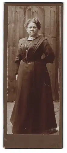 Fotografie unbekannter Fotograf und Ort, ältere Dame im dunklen Kleid mit Halskette vor einer Holzwand