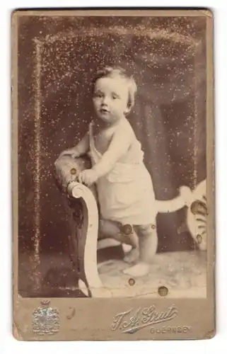 Fotografie T. A. Grut, Guernsey, Süsses Kleinkind im Hemd steht auf Sofa