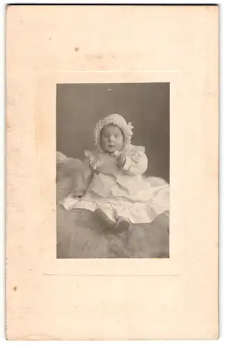 Fotografie unbekannter Fotograf und Ort, Kleinkind im weissen Kleidchen mit Haube sitzt auf einem Fell
