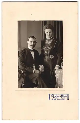 Fotografie Esch & Stein, Mülheim / Ruhr, Paar im feinen Anzug mit Fliege und im Kleid mit Halskette, toupierte Haare
